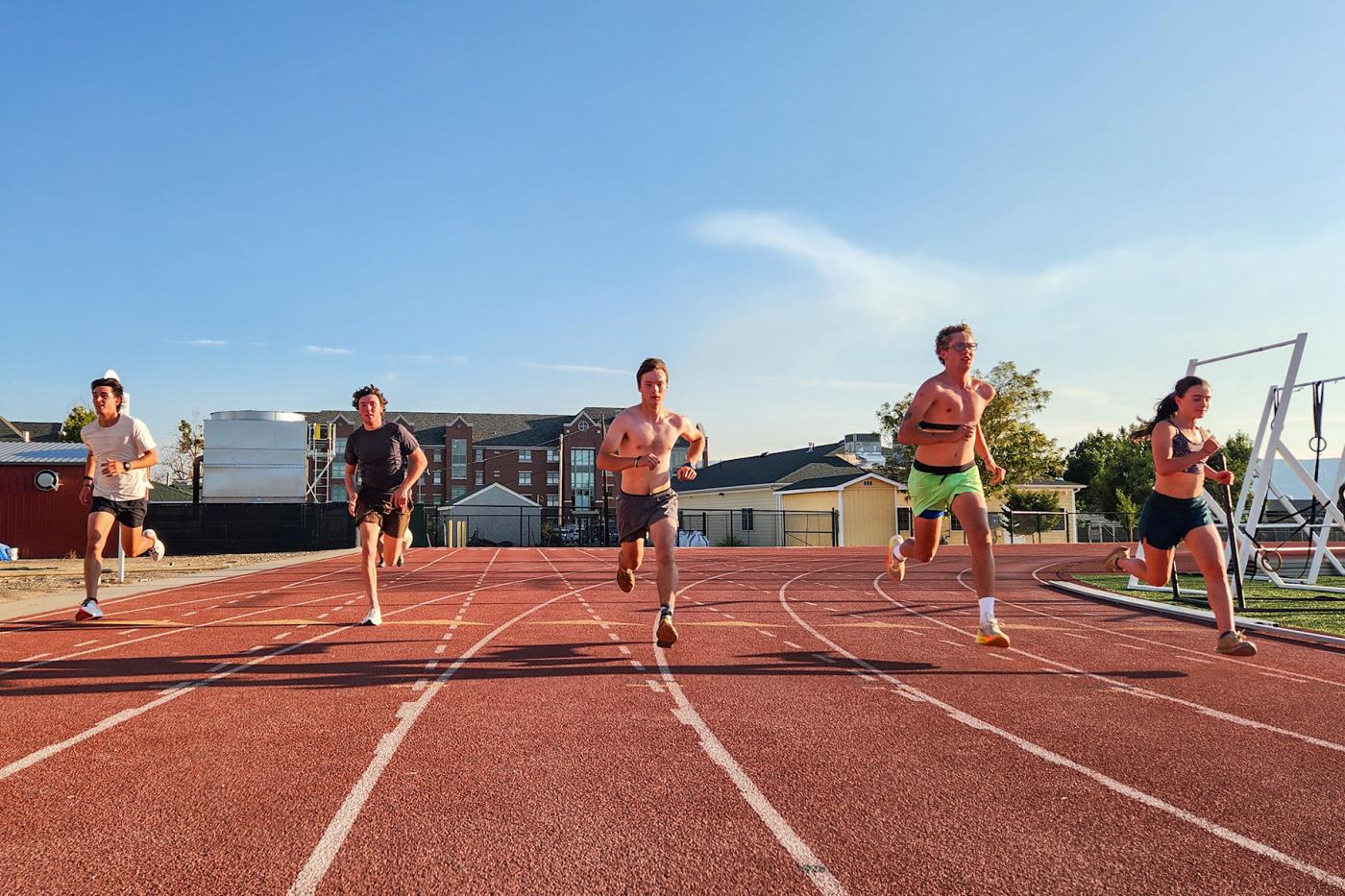 Five college athletes sprinting on an outdoor track in the evening sun during a training session.